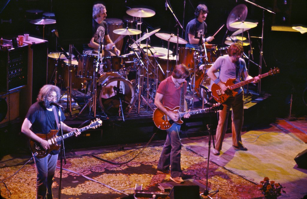 Image Description: The Grateful Dead at the Warfield Theatre in San Francisco, October 9, 1980 from Chris Stone. Features three men on a stage playing guitars, and two men in the background playing drums. Image licensed for reuse through Wikimedia Commons. 