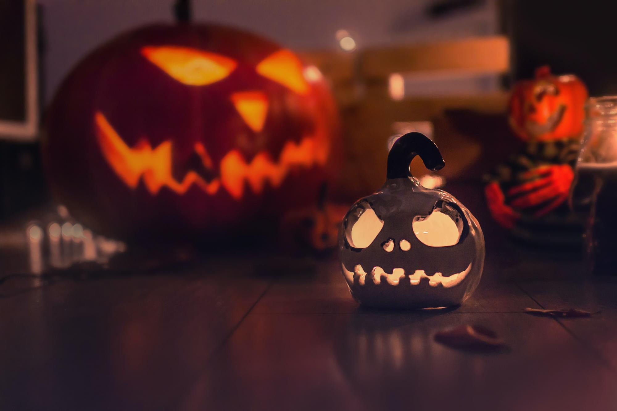 A small whitish pumpkin with a sinister face carved into it sits in the foreground of the image, with a much larger orange pumpkin with a similar face carved into it sitting in the background.