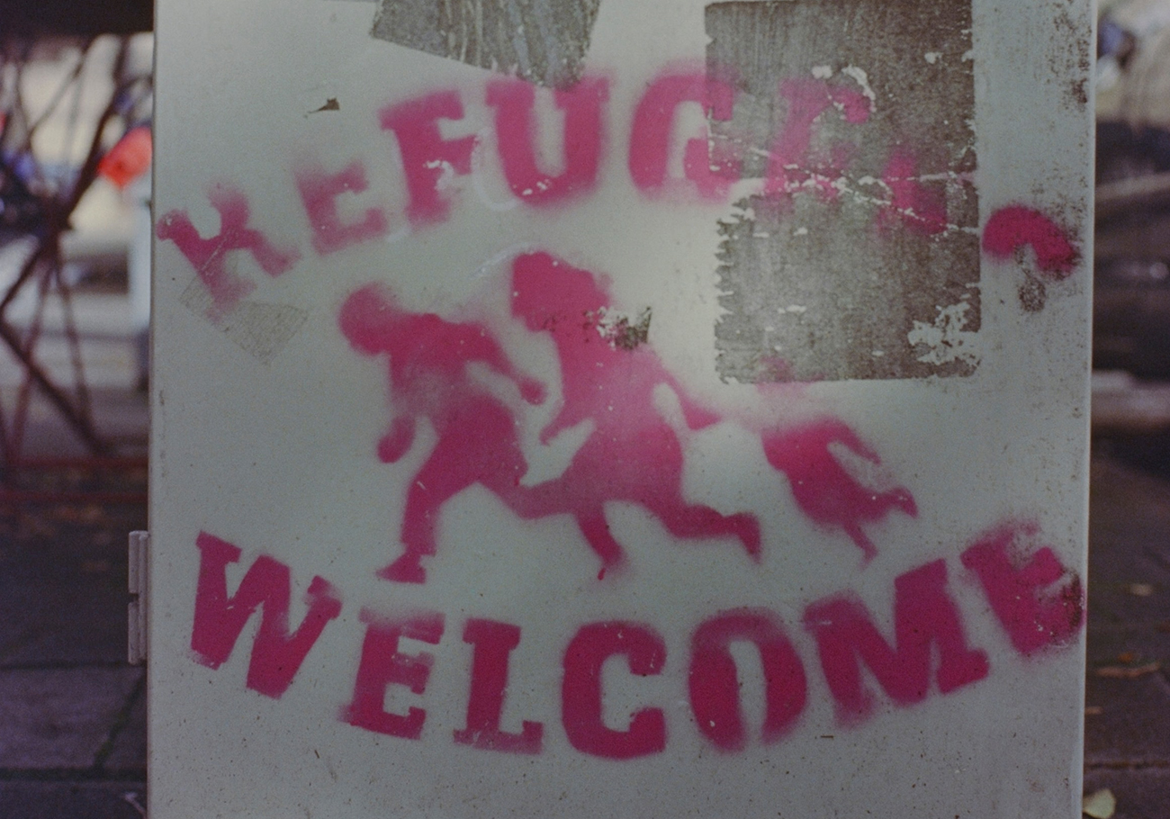 Image of stenciled graffiti of "Refugees Welcome" with a silhouette of a family running to safety.