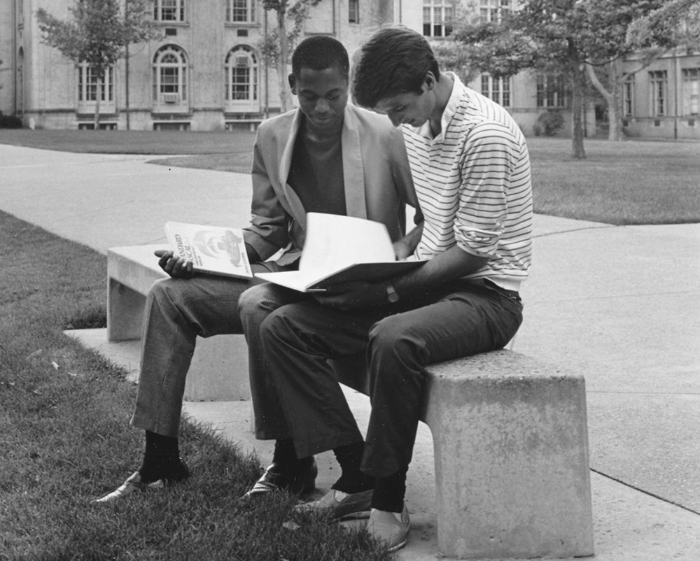 Two college students sit on a bench studying. One student holds a copy of Doug Cooper's "Standard Pascal User Reference Manual." Baker Hall and part of The Mall can be seen in the background.