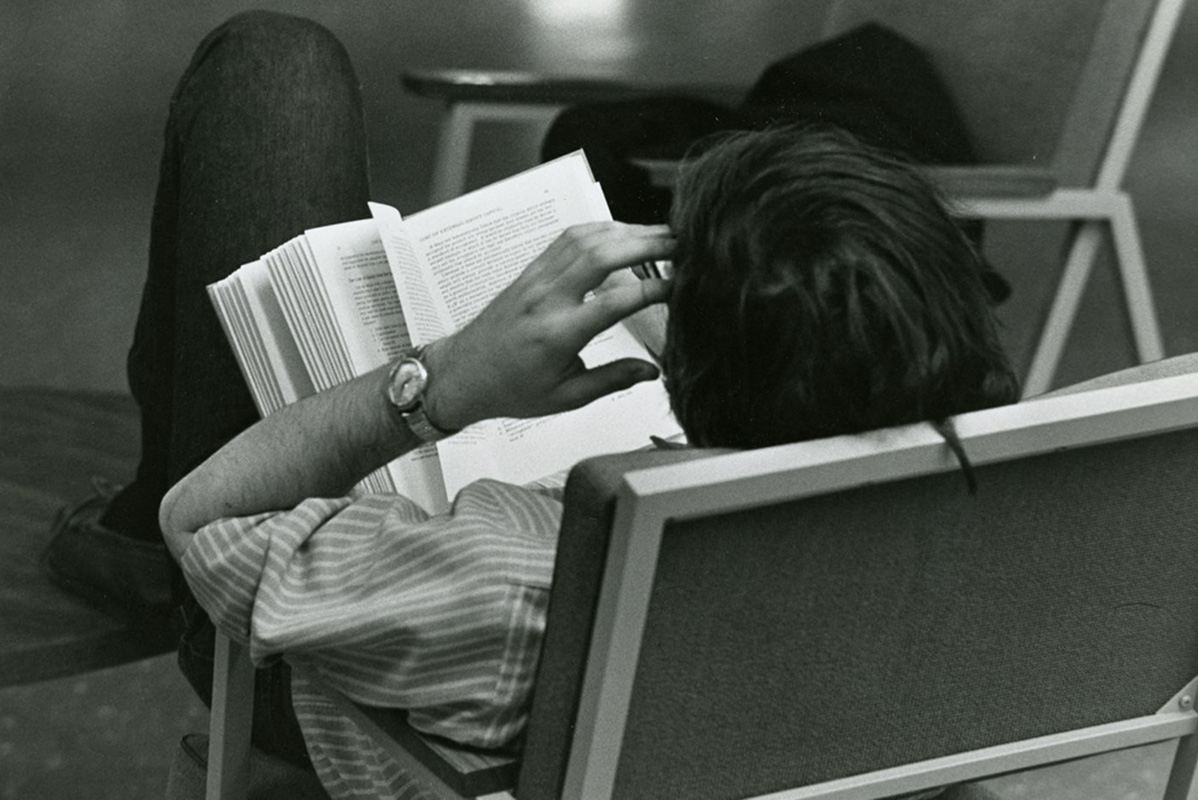 A college student sits back in a chair and reads a book. (c.1985)