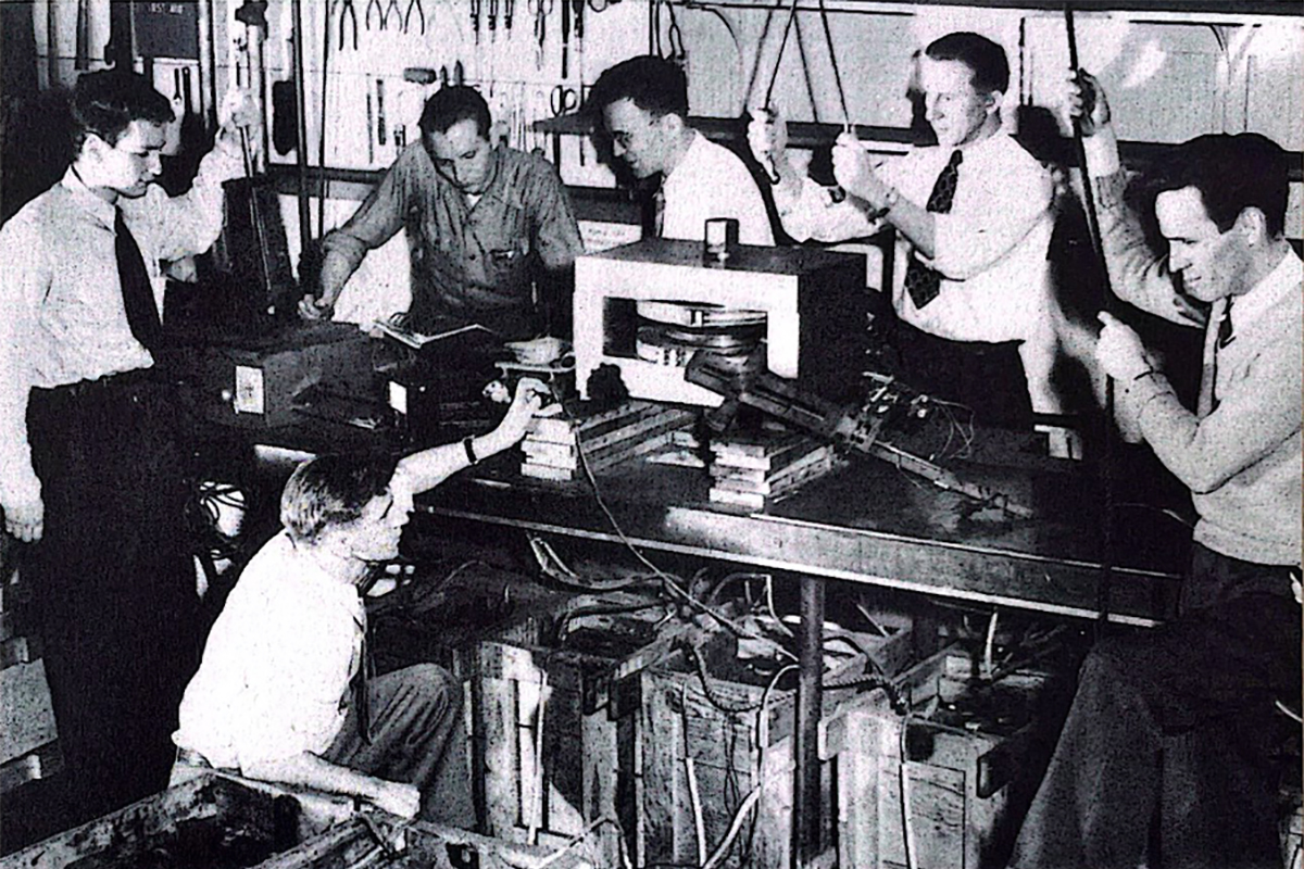 Scientists at Carnegie Tech experiment with a small model of the cyclotron. The storage batteries in the wooden boxes under the table are capable of furnishing up to five thousand amperes for exciting the model coils. (Left to right) Rolf Winter, Ray Sedney (stooping), Irving Zacher, Dr. Martyn Foss, James Mates, Dr. John Fox.